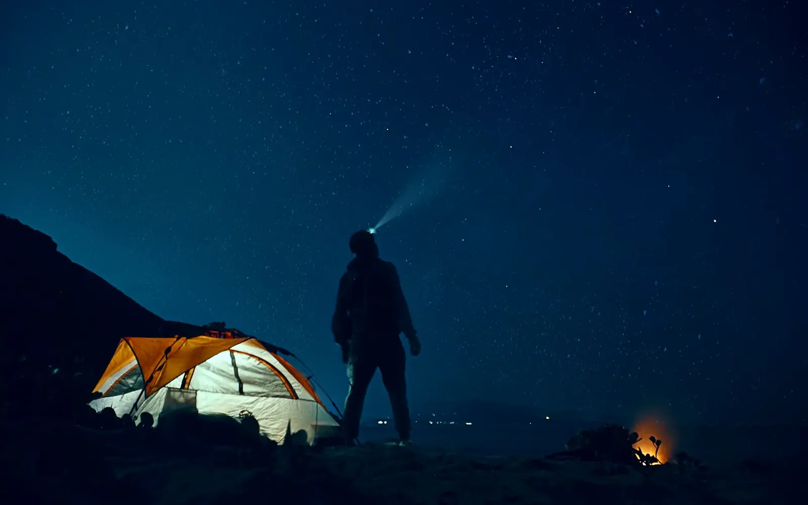 Persona con linterna frontal encendida de noche junto a una carpa bajo las estrellas