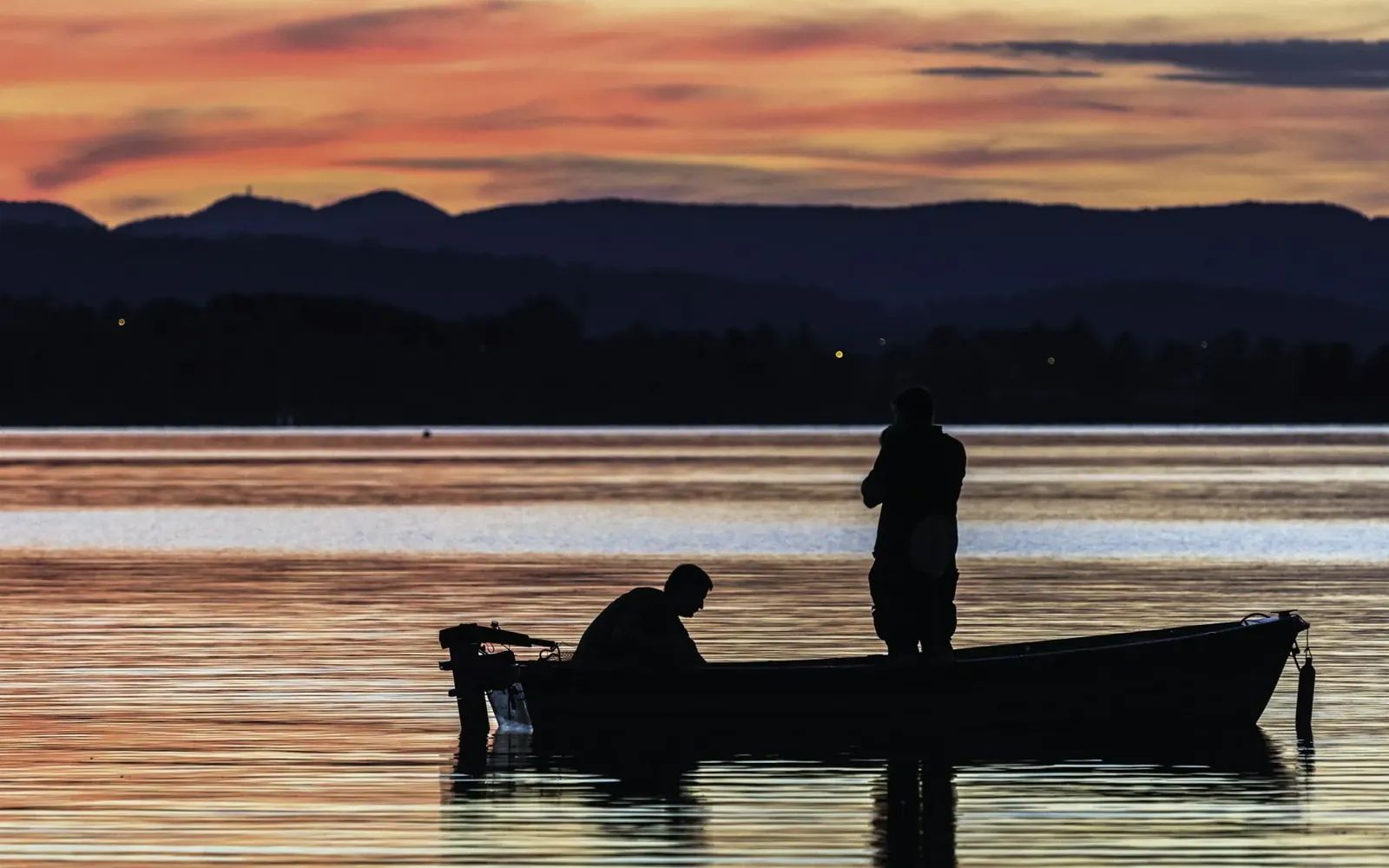 Dos pescadores en un bote sobre un lago al atardecer, con sierras de fondo